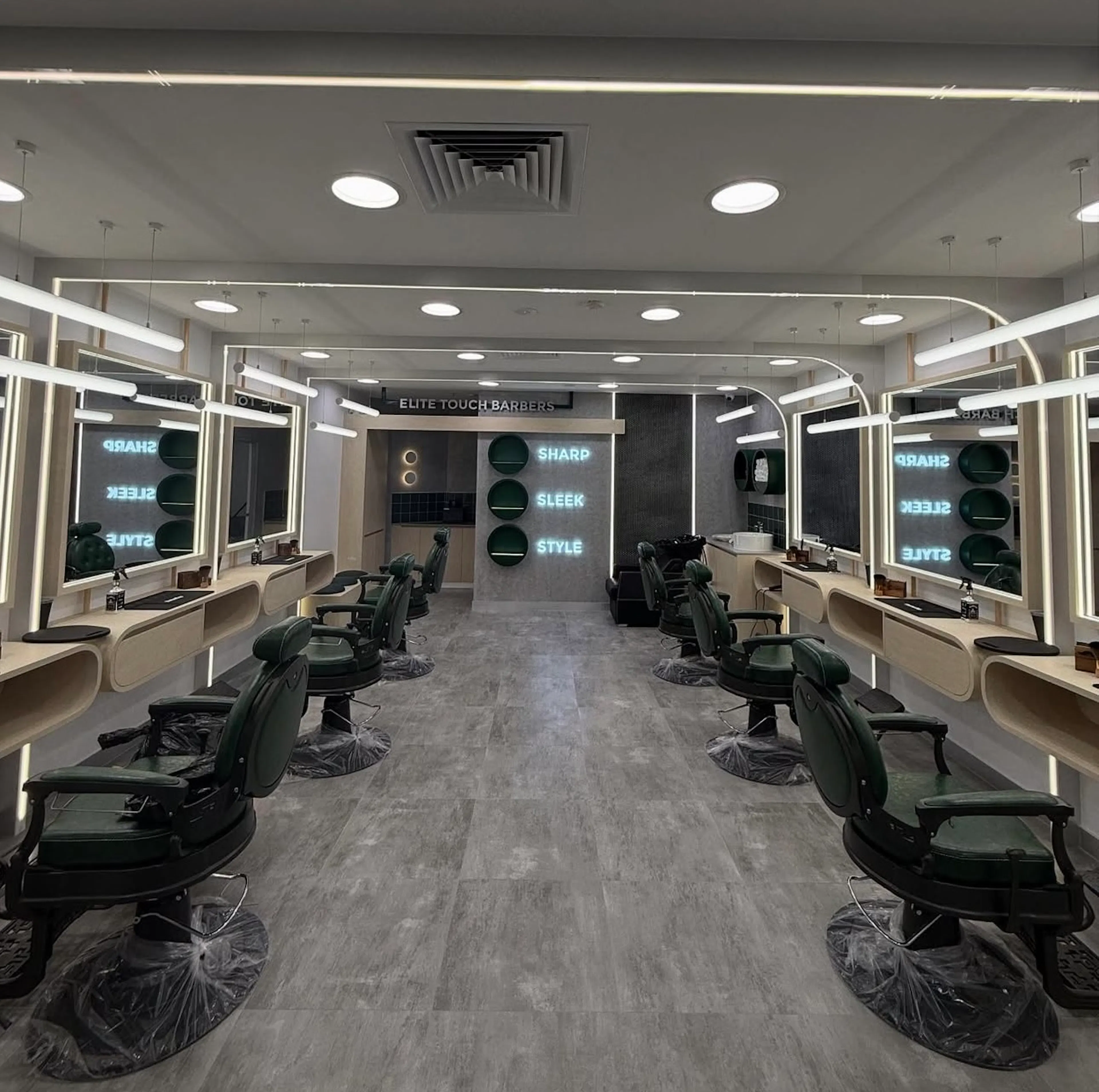 Central aisle view of a modern barbershop flanked by backlit mirror stations and green chairs.
