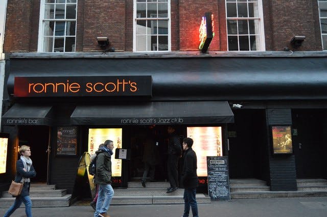 A photograph of "Ronnie Scott's" jazz club, as seen from the other side of the road on which it is located. The front of the club is painted black, with black awnings covering the doors. The sign over the door is black, with lowercase orange lettering reading "ronnie scott's". 