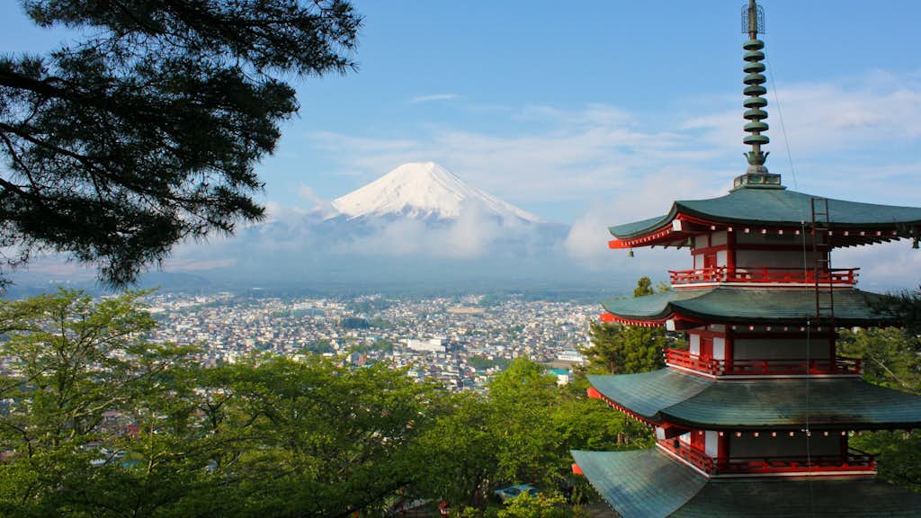 Eine malerische Ansicht des schneebedeckten Berges Fuji im Hintergrund, mit einem traditionellen, mehrstöckigen, roten Pagodenturm im Vordergrund. Die Szene ist von grünen Bäumen eingerahmt und bietet einen Blick über die Stadt, die sich in der weiten Ebene unterhalb des Berges erstreckt.