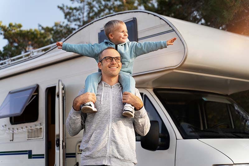 Men with child on shoulders standing in front of caravan