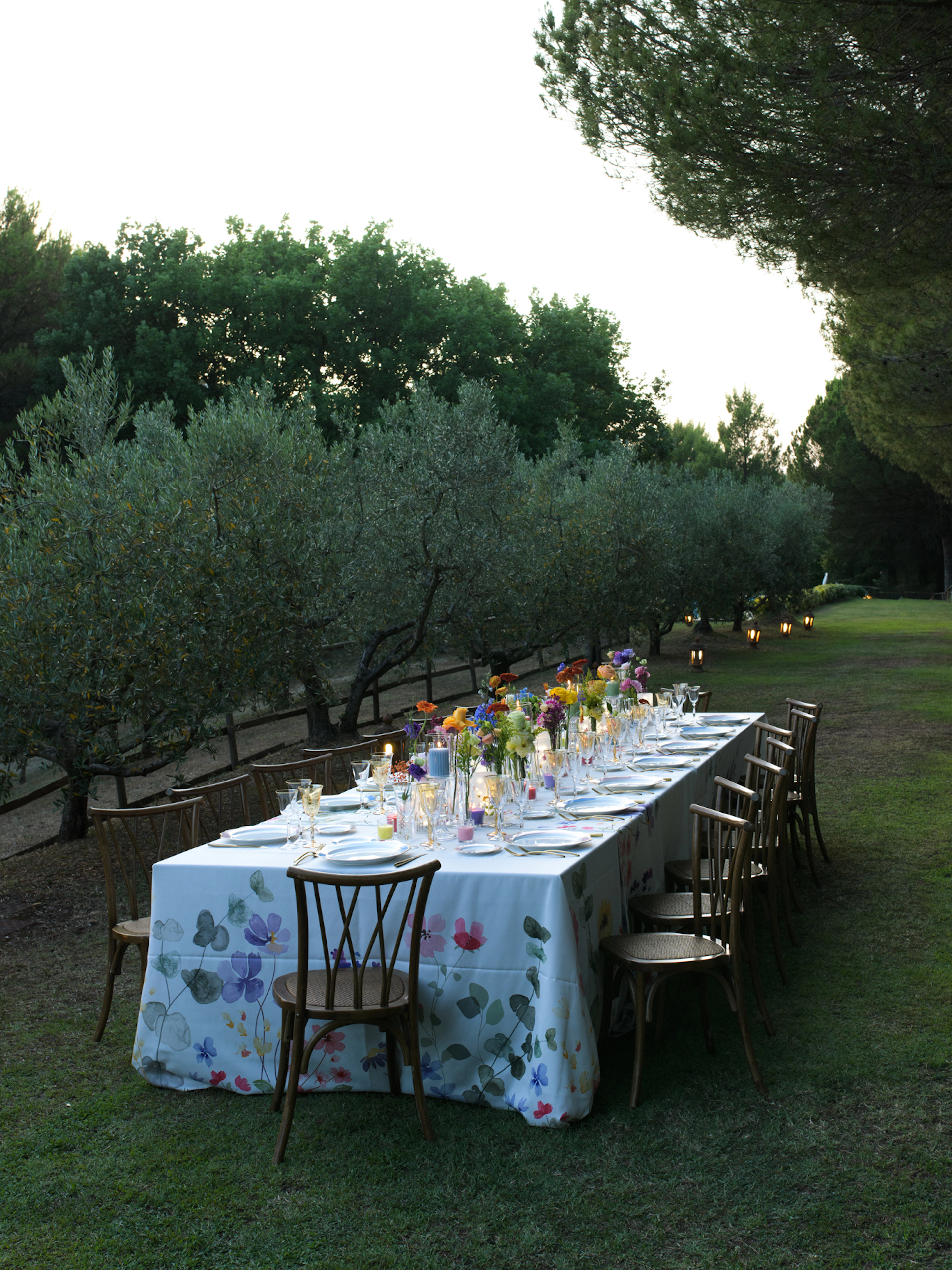 Intimate outdoor dining table with crystal chandelier, rattan chairs and white floral centerpiece at dusk at La Fonte estate, Italy