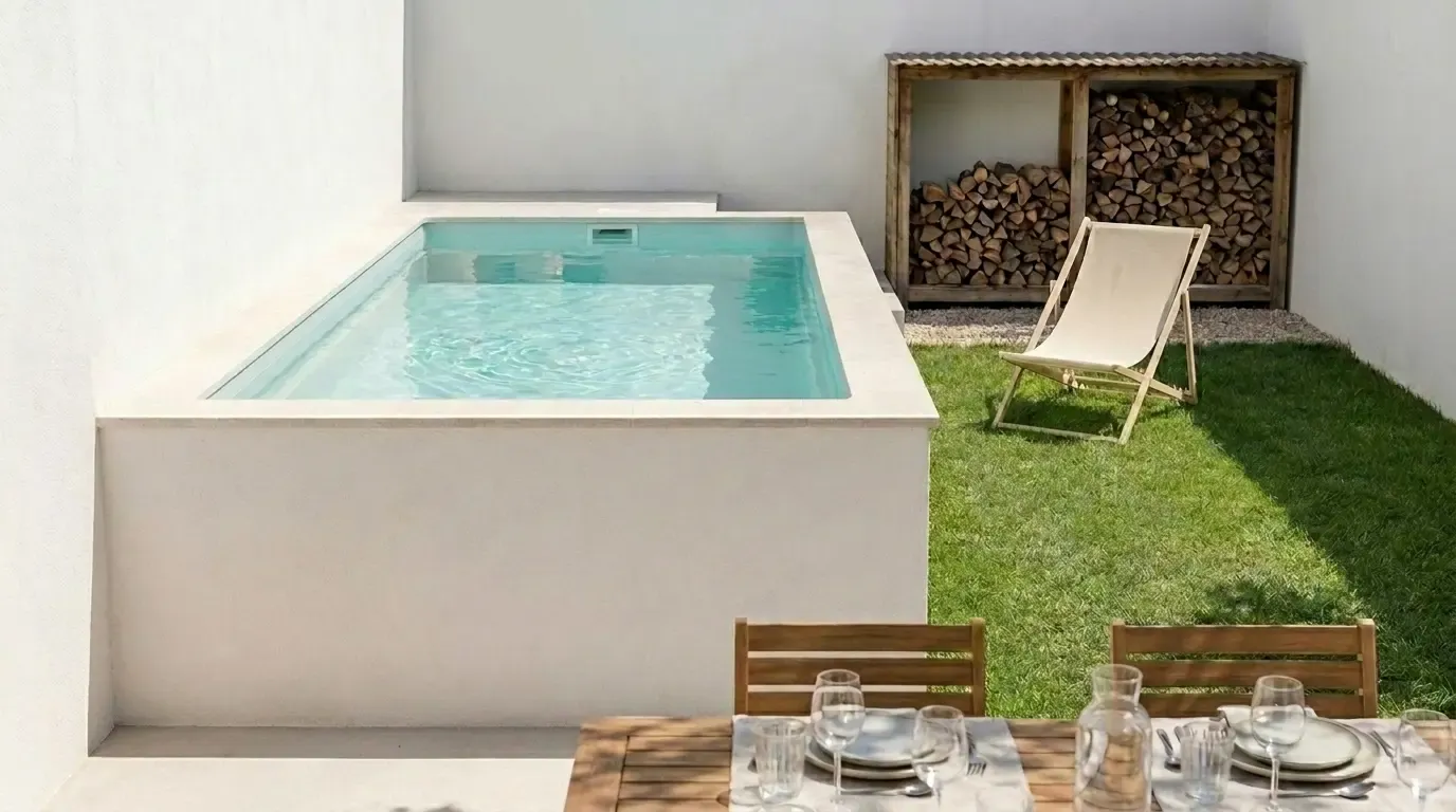 Mini-piscine surélevée habillée d'un enduit clair, abri à bois et transat en toile beige sur pelouse