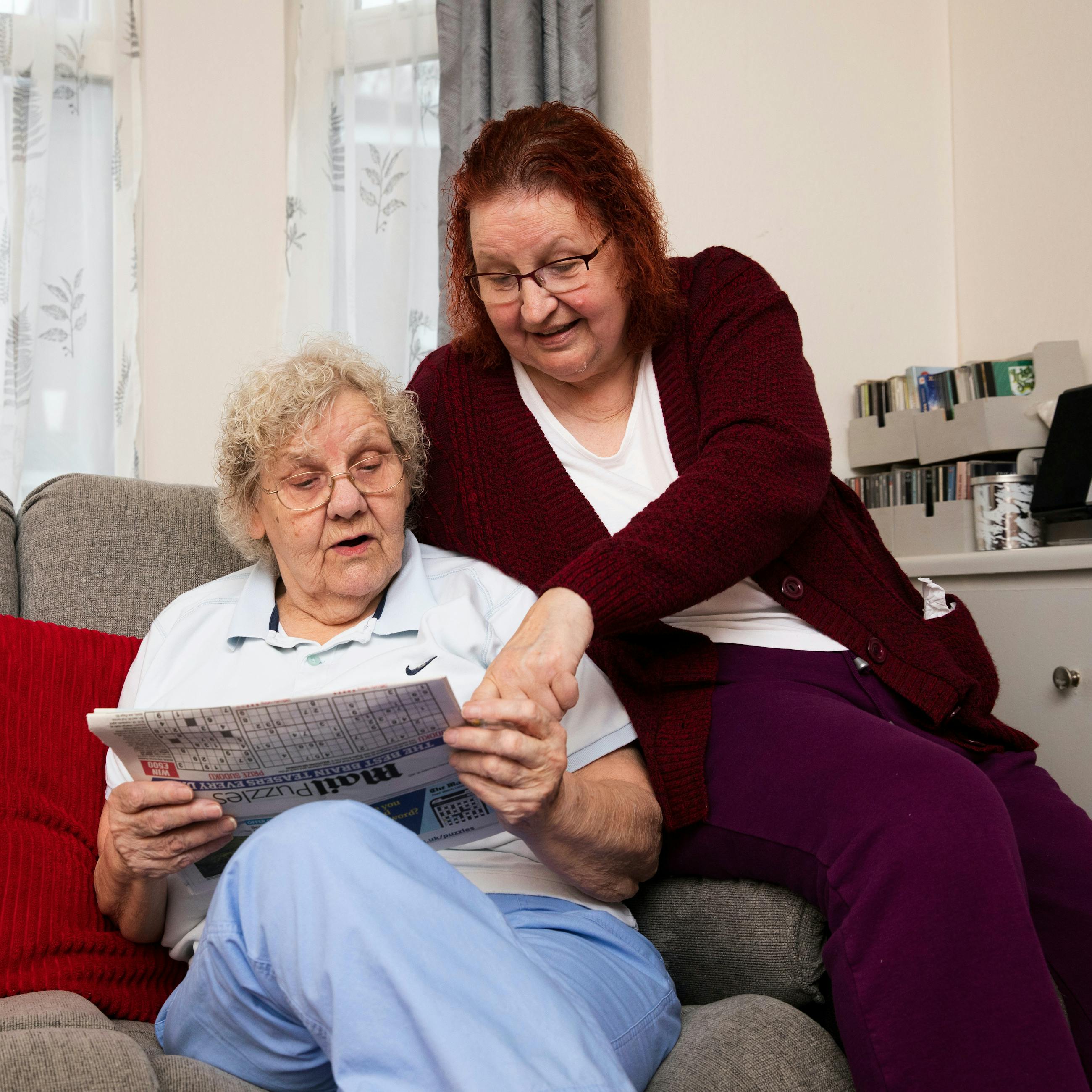 Two older women reading a newspaper at home, one pointing at an article.