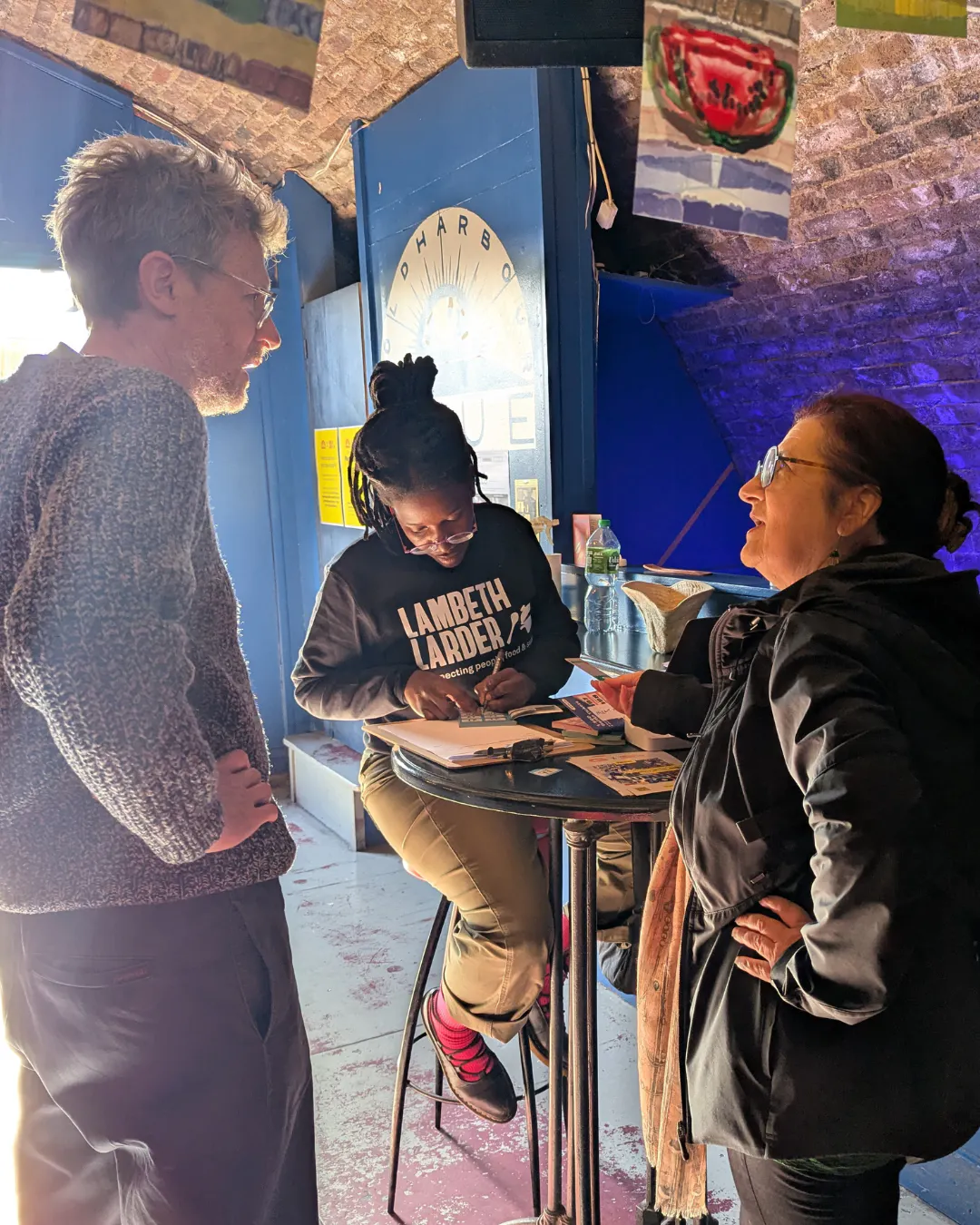 Two people chatting in front of a woman with a Lambeth Larder sweat shirt who is writing something on a piece of paper.