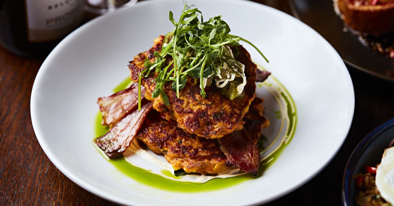 A vibrant plate of Lantana's signature corn fritters, stacked with fresh spinach, roasted tomatoes, smashed avocado, and topped with lemon crème fraîche, served with a side of bacon.