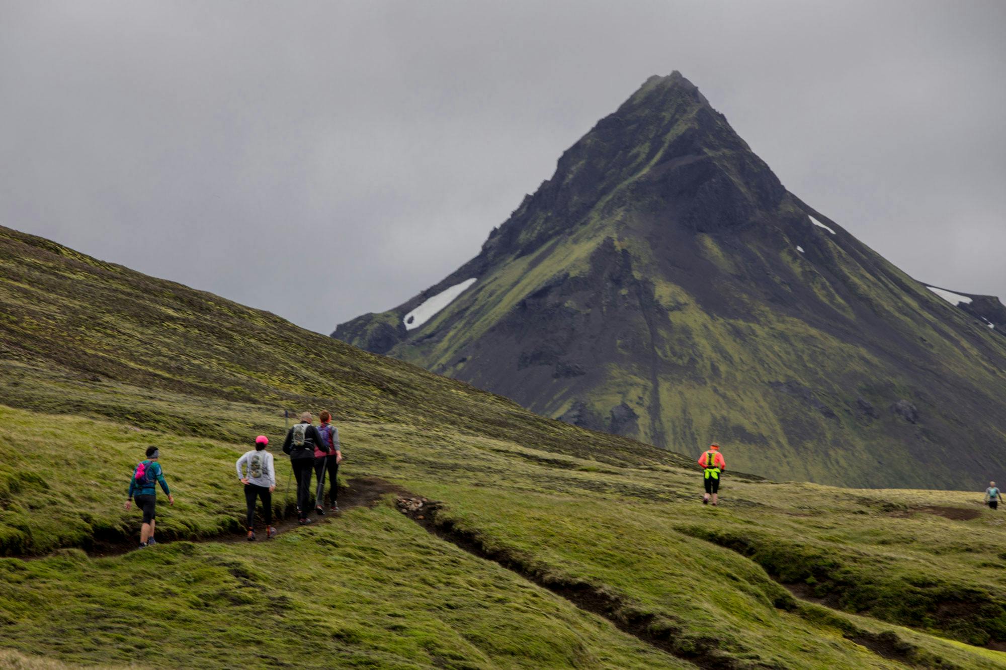 Few runners running in green landscape towards a black mountain