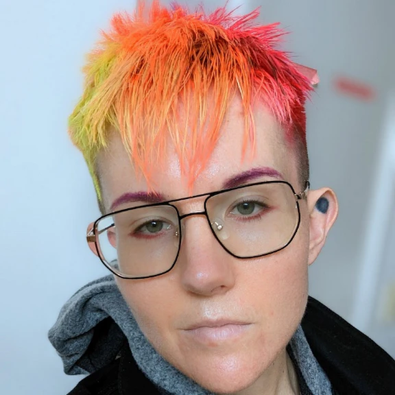 Photo of a non-binary person with short orange-pink-yellow gradient hair and aviator glasses.