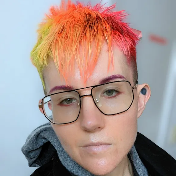 Photo of a non-binary person with short orange-pink-yellow gradient hair and aviator glasses.
