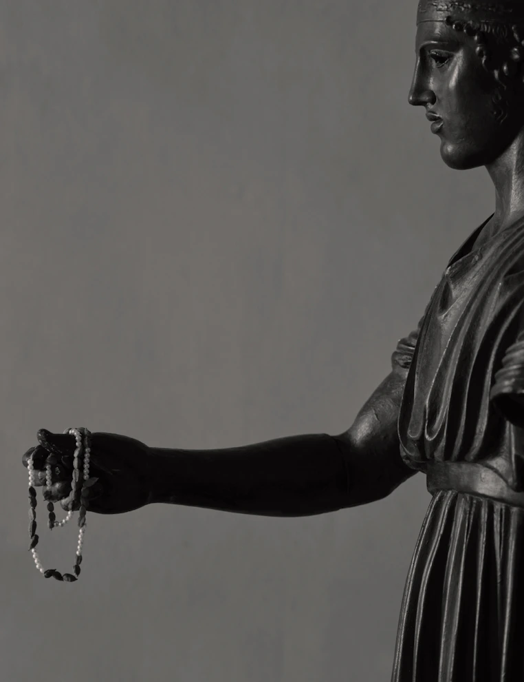 Side profile of the Charioteer of Delphi bronze statue holding reins against a plain background.