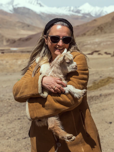 Mariella Ienna wearing a sunglasses and a tan shearling jacket cradling a baby goat in a dry valley with snow-capped mountains in the distance.