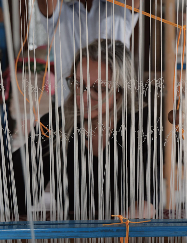 Mariella seen through dense vertical warp threads, face partially obscured by the loom.