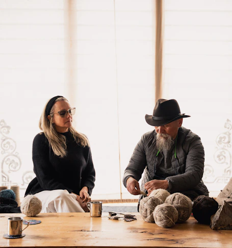 Founder with a man sitting at a light wooden table with yarn, talking and working with thread.