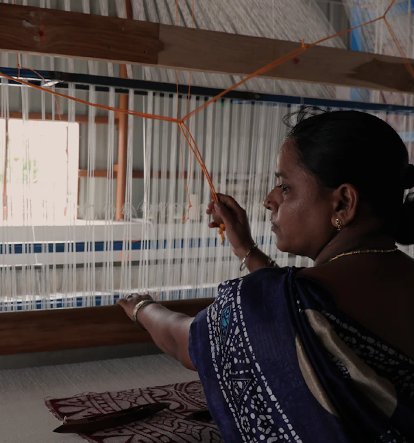 Woman working a tall wooden handloom, surrounded by dense vertical white threads.