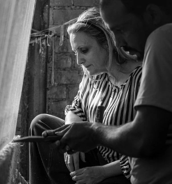 Founder with a man working together at a loom, focused on the threads in a brick walled workshop.
