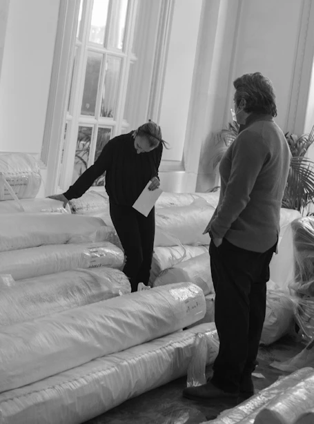 Two people at Grand Hotel examining large stacked rolls of rugs wrapped in plastic.