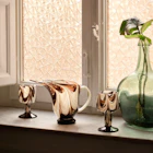 Swirled amber glass pitcher and goblets beside a green vase on a marble windowsill.