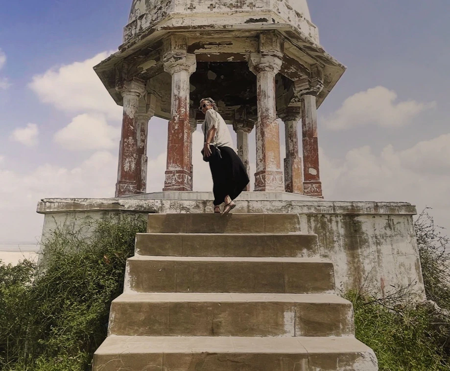 Woman standing inside a weathered stone pavilion on a raised platform, looking out over a hazy landscape under a pale blue sky with clouds.