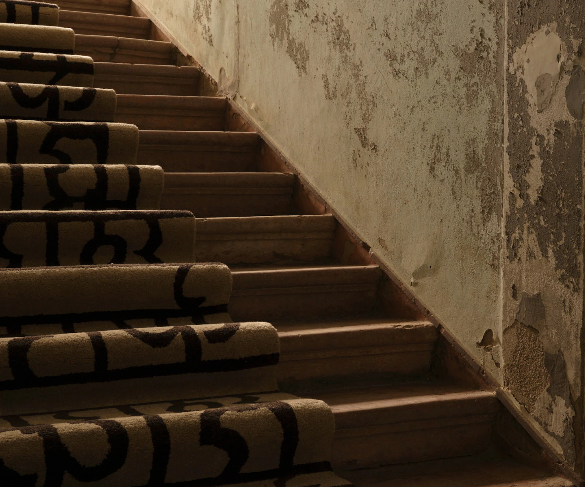 A dim, atmospheric photograph of a staircase with a thick, light‑brown Lavor Ombra rug on the lower steps, patterned with bold, dark calligraphic markings.