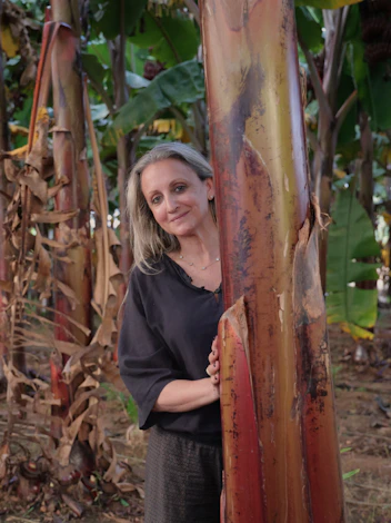 Mariella standing in a banana grove, partially hidden behind a textured banana tree trunk.