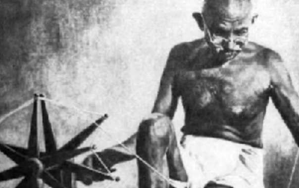 Mahatma Gandhi seated on the floor operating a spinning wheel, drawing a fine thread by hand.