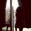 Silhouetted figure standing in a dark interior doorway, looking out over sunlit Palermo rooftops under a bright sky.