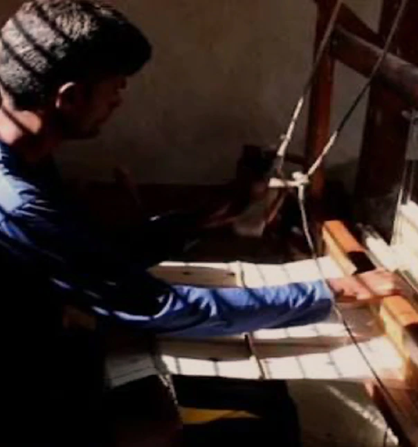 Young man in side profile operating a handloom, strong light highlighting striped white fabric.