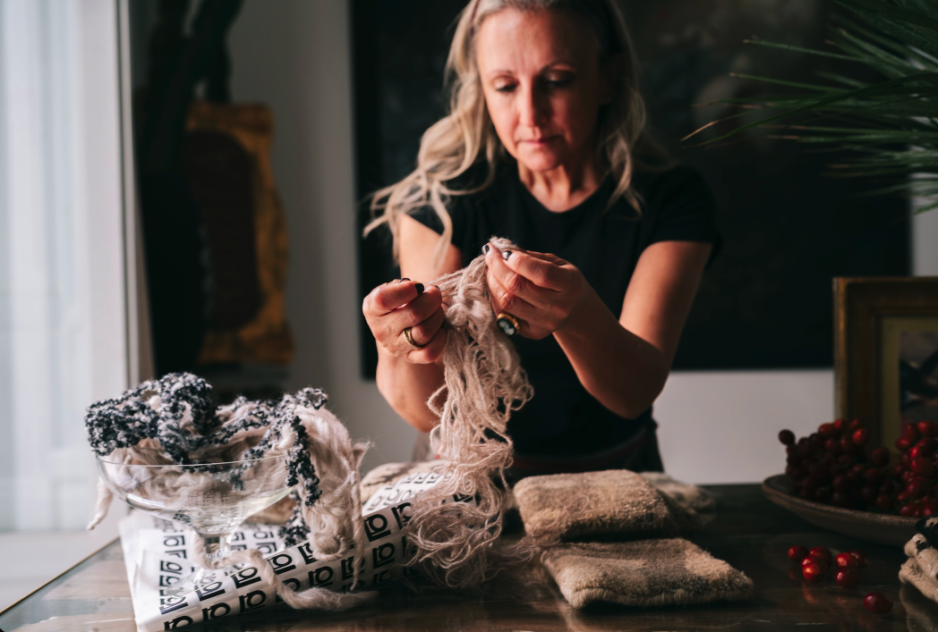 A photograph of the founder, Mariella Ienna  seated at a table in a dimly lit room, focused on working with bundles of pale fibrous material laid out before her.