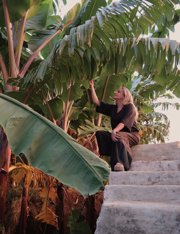 Mariella, the founder sitting on outdoor steps under large banana leaves, reaching up to touch the foliage.
