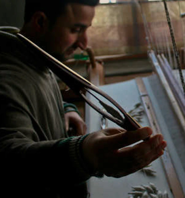 Bearded man at a large wooden loom guiding a boat shuttle through pale warp threads.