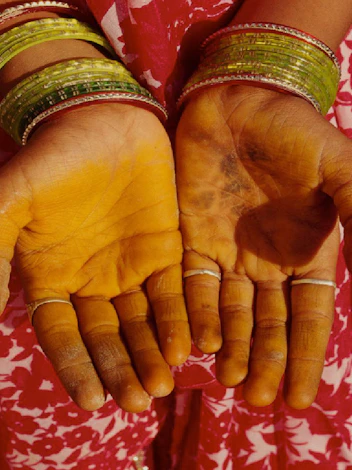 Open palms stained bright yellow with turmeric, framed by green glass bangles on a red patterned saree.