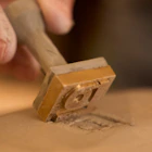 Hand pressing a metal stamp with a circular logo into a smooth tan surface.