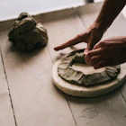 Hands shaping a dark clay ring on a light wooden table, with a lump of clay nearby.