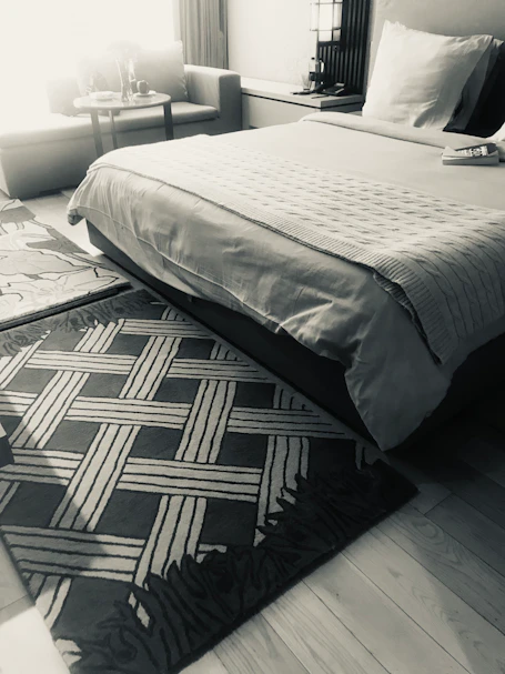 Modern bedroom with a geometric dark rug beside a neatly made light-textured bed.