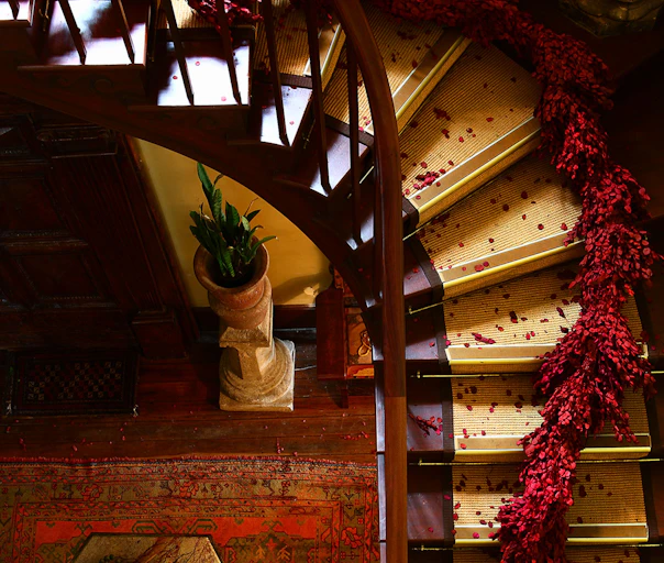Top-down view of a grand dark-wood staircase with pale carpet runners, scattered red petals, and a cascading red floral garland, curving above a patterned rug and potted green plant on a pedestal in a moody, antique interior.