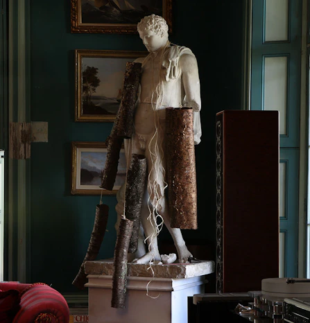 White marble male statue indoors, wrapped with rough bark-like forms and trailing white fibers, standing before dark teal walls with framed landscape paintings and vintage audio equipment.