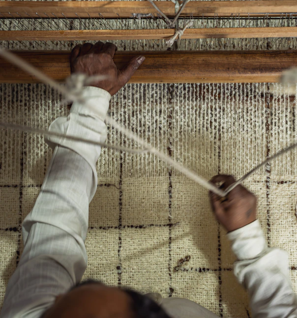 Overhead view of a weaver’s hands working a cream rug with a dark grid pattern on a loom.