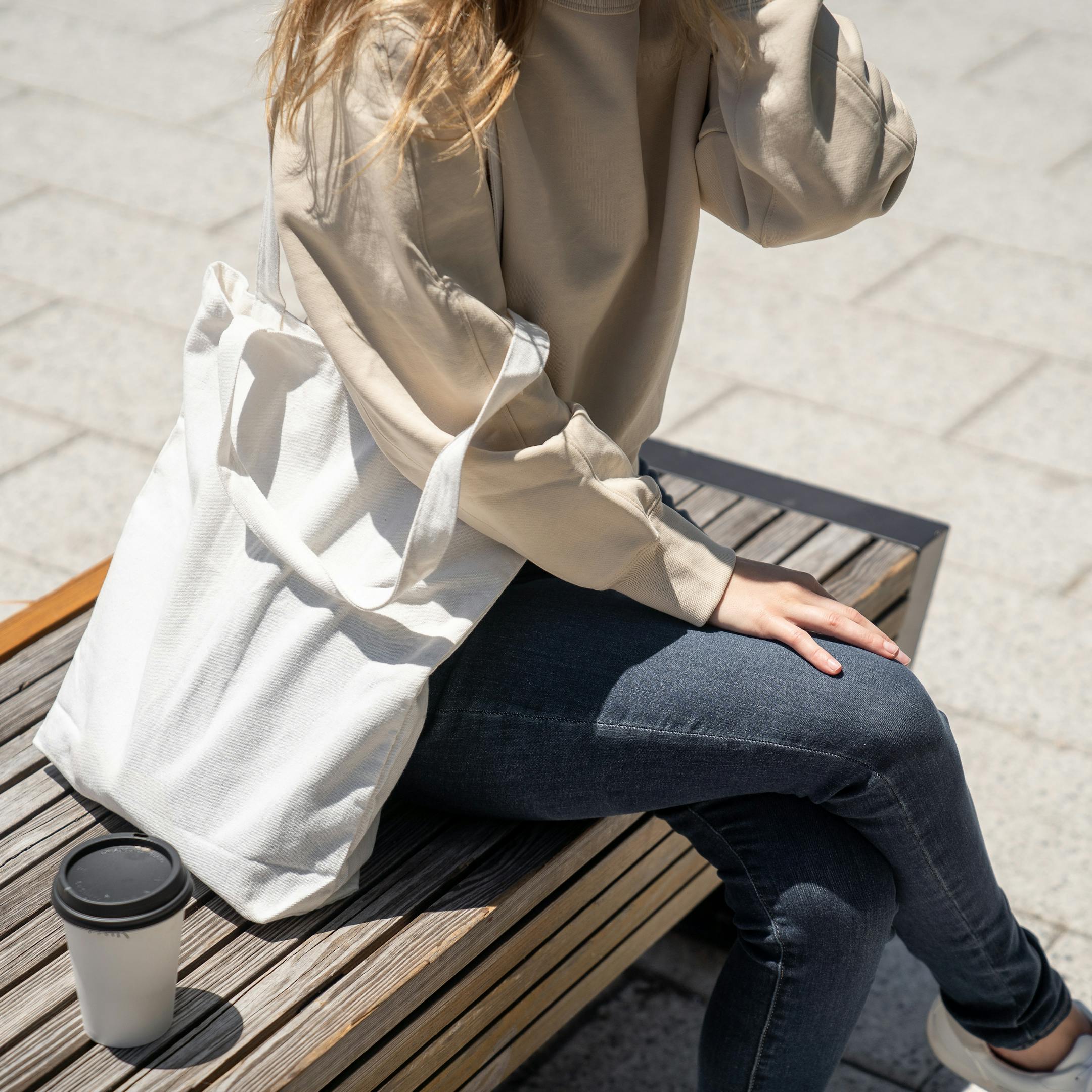 Femme assise sur un banc avec un café et un sac en toile blanc