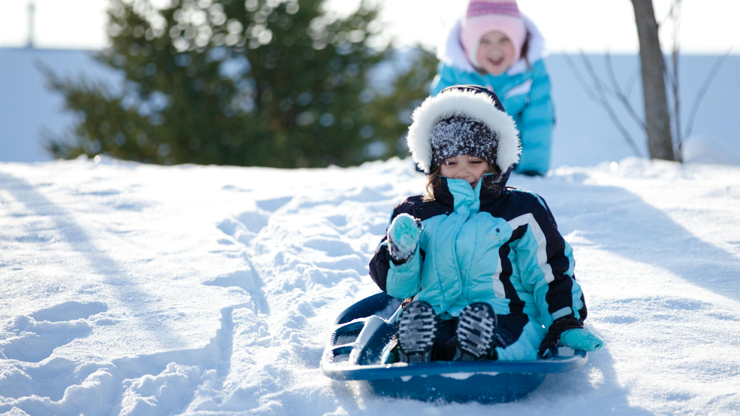 Deux petites filles faisant de la luge dans la neige, l'une pousse l'autre. 