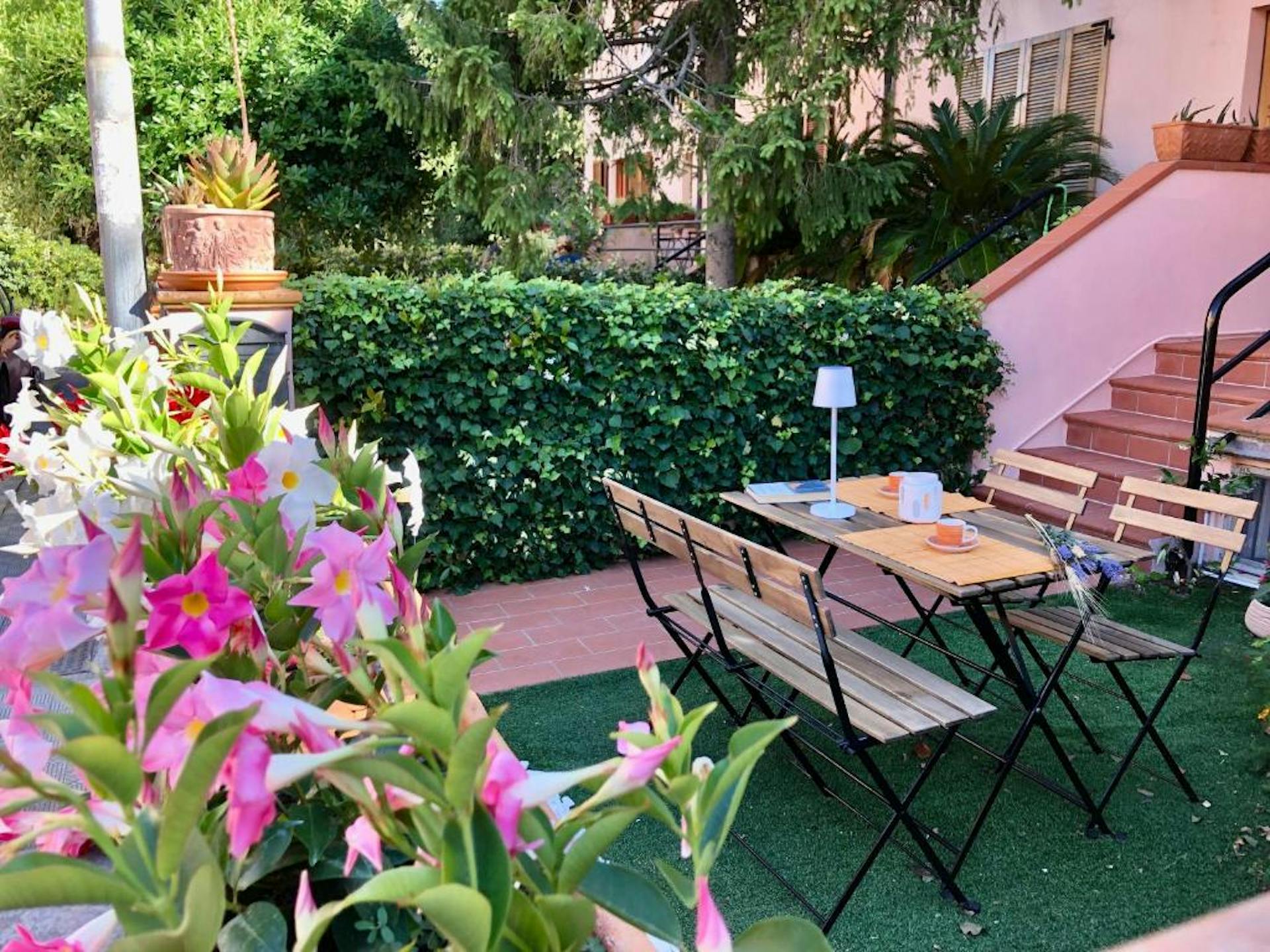 Dettaglio del tavolo esterno con quattro sedie in legno e fiori sul rosa