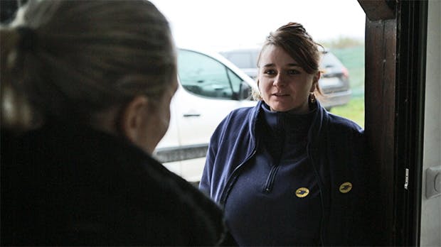 Photo prise de l'intérieur d'un domicile avec une factrice saluant la résidente (vue de dos) sur le pas de sa porte.