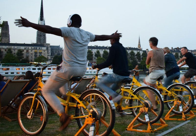 Photo de personnes qui pédalent sur des vélos jaunes face à l'écran (hors champ) d'un cinéma en plein air.