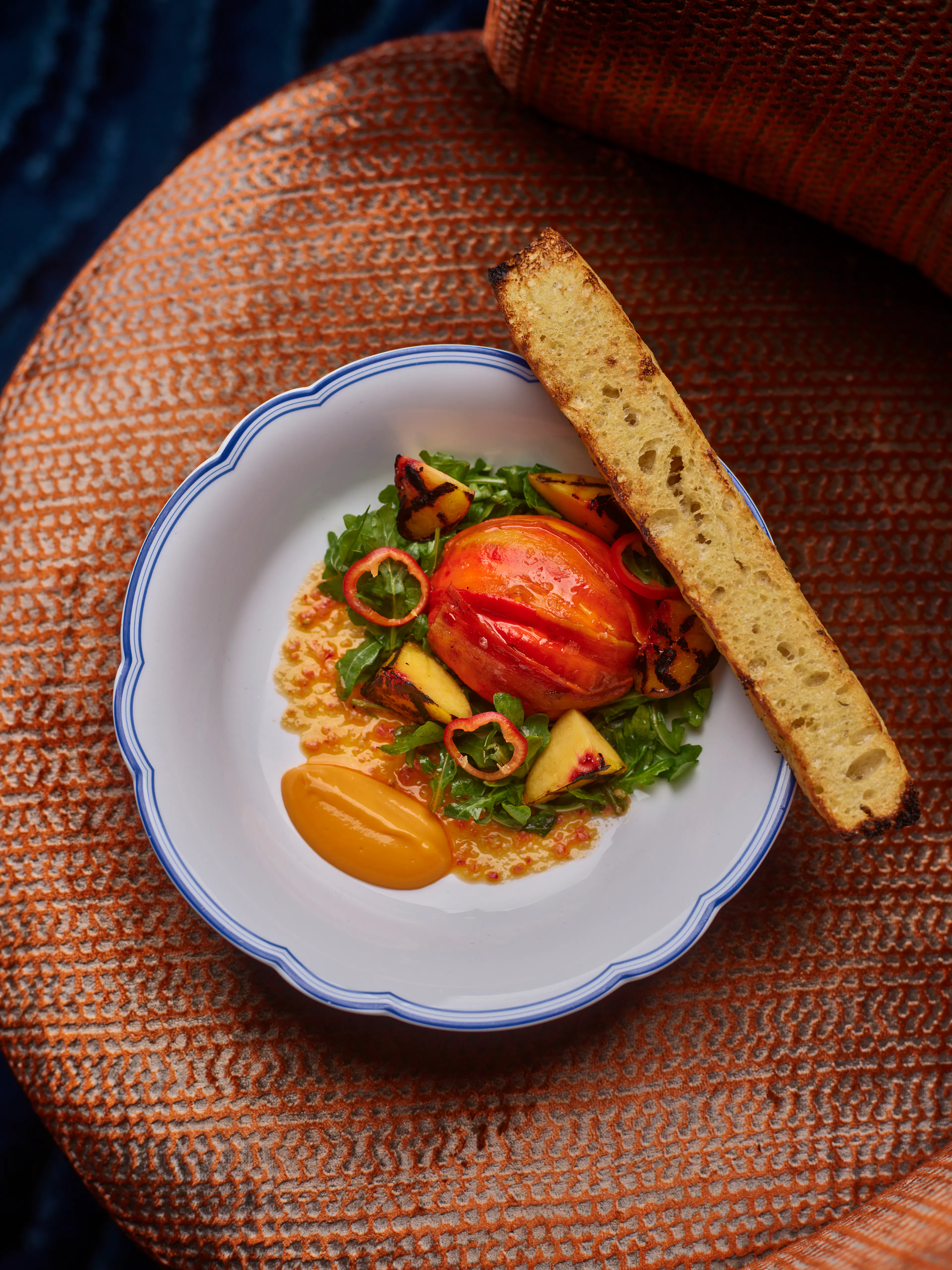 Summer Burrata with Jimmy Nardello peppers, grilled peaches, fresh greens, and toasted focaccia, served in a white and blue-rimmed bowl.