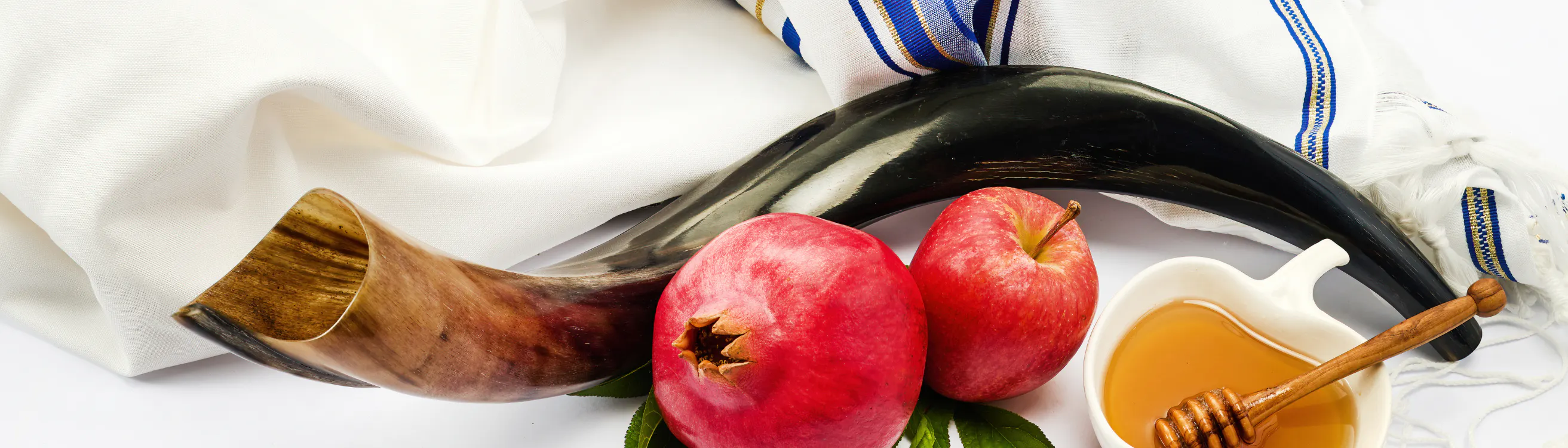 Shofar, pomegranate, apple, and a heart-shaped bowl of honey arranged on a white cloth with a blue-striped tallit for Rosh Hashanah.