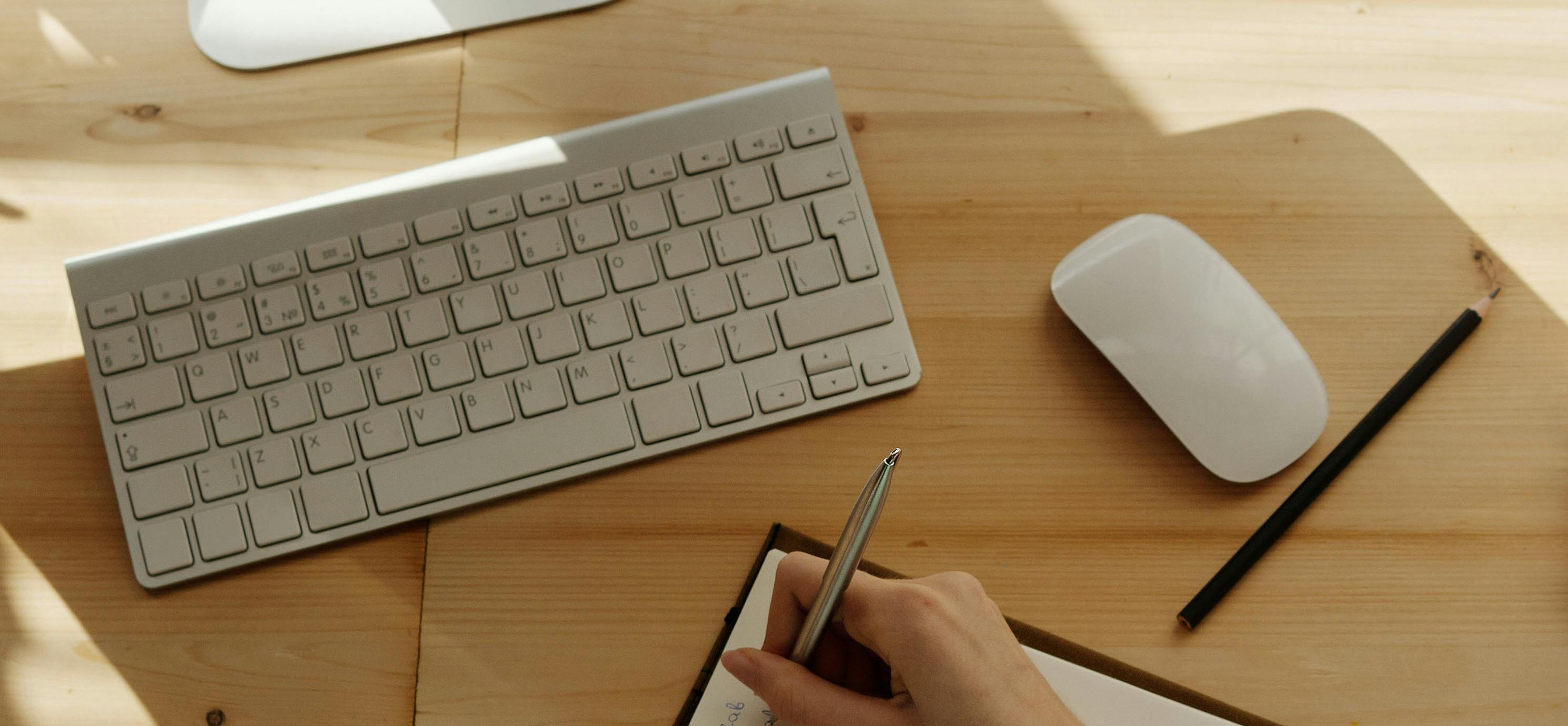 Close-up of a desk with a wireless keyboard, wireless mouse and a hand writing notes on a piece of paper