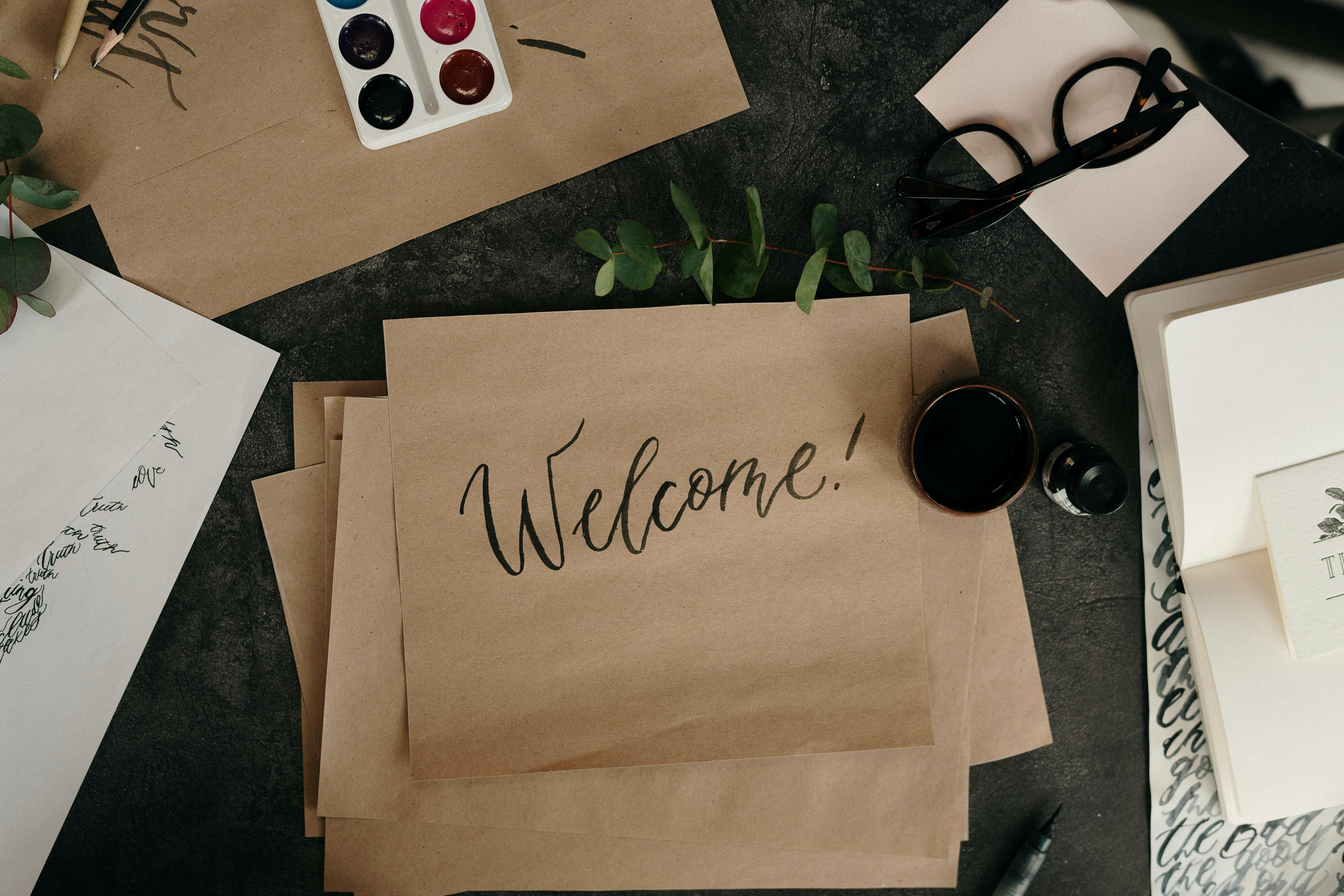 Stack of paper and paints on a table, one piece of paper has the word "Welcome" written on it.