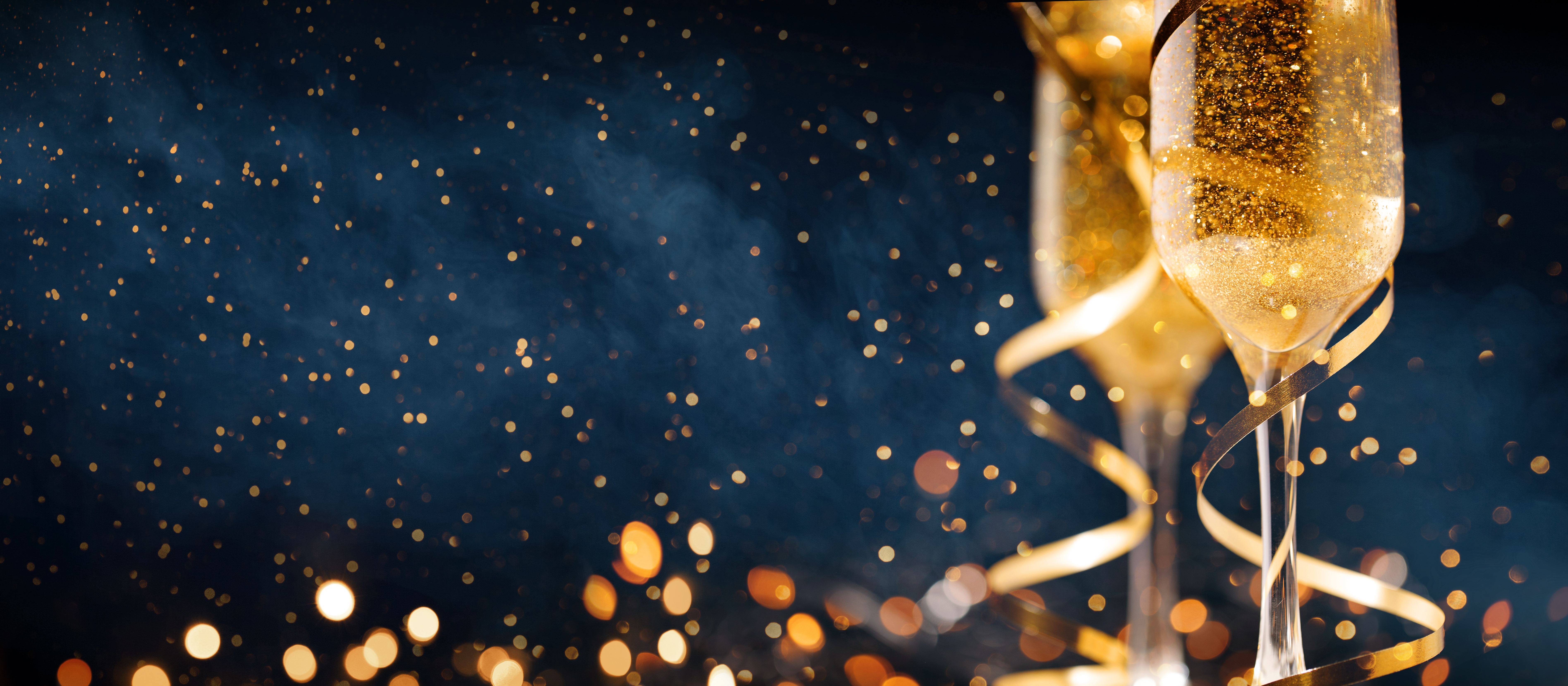 Two champagne glasses filled with golden sparkling liquid, adorned with ribbons, set against a festive backdrop of twinkling lights.
