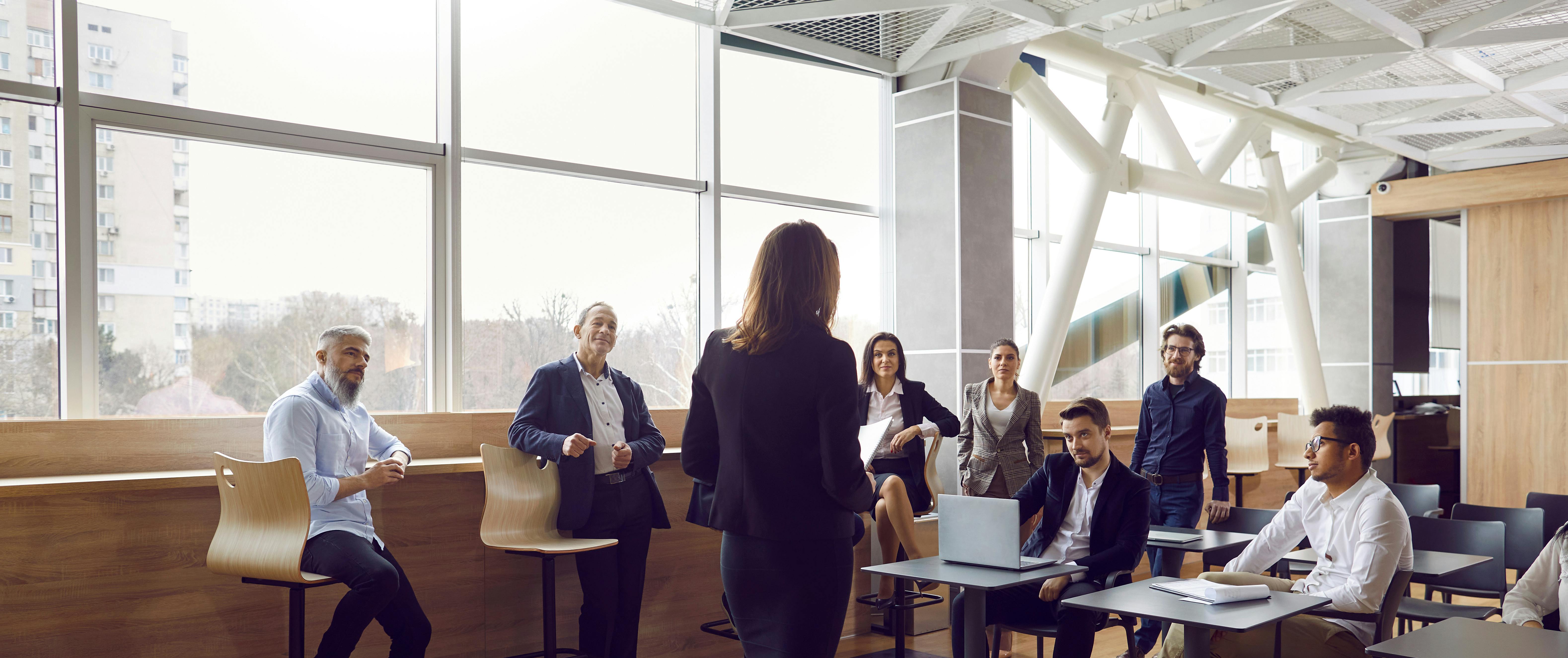 A woman addresses a group of attentive professionals seated in a bright, open space.