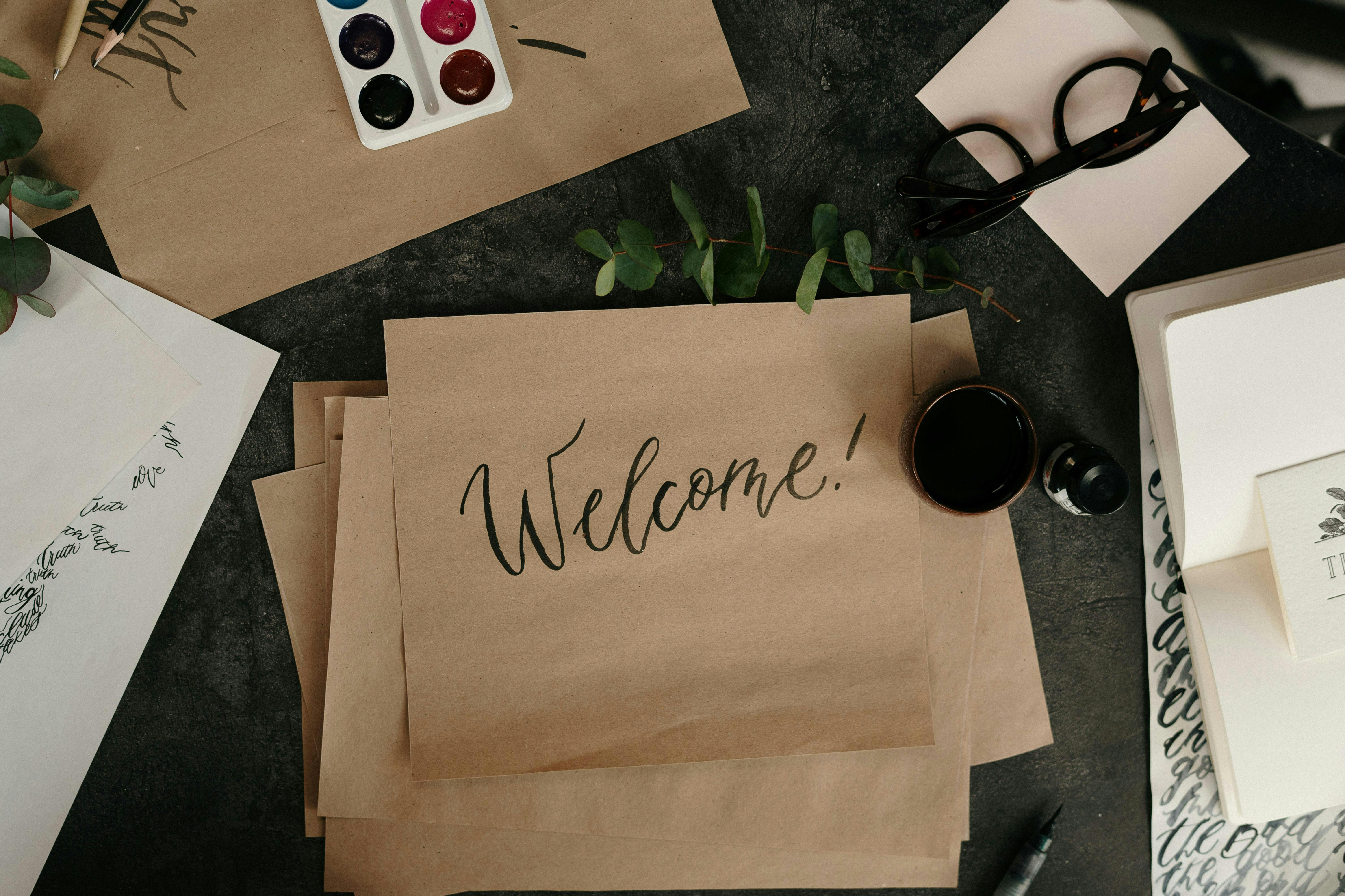 Handwritten "Welcome!" on brown paper, surrounded by calligraphy tools, glasses, and greenery.