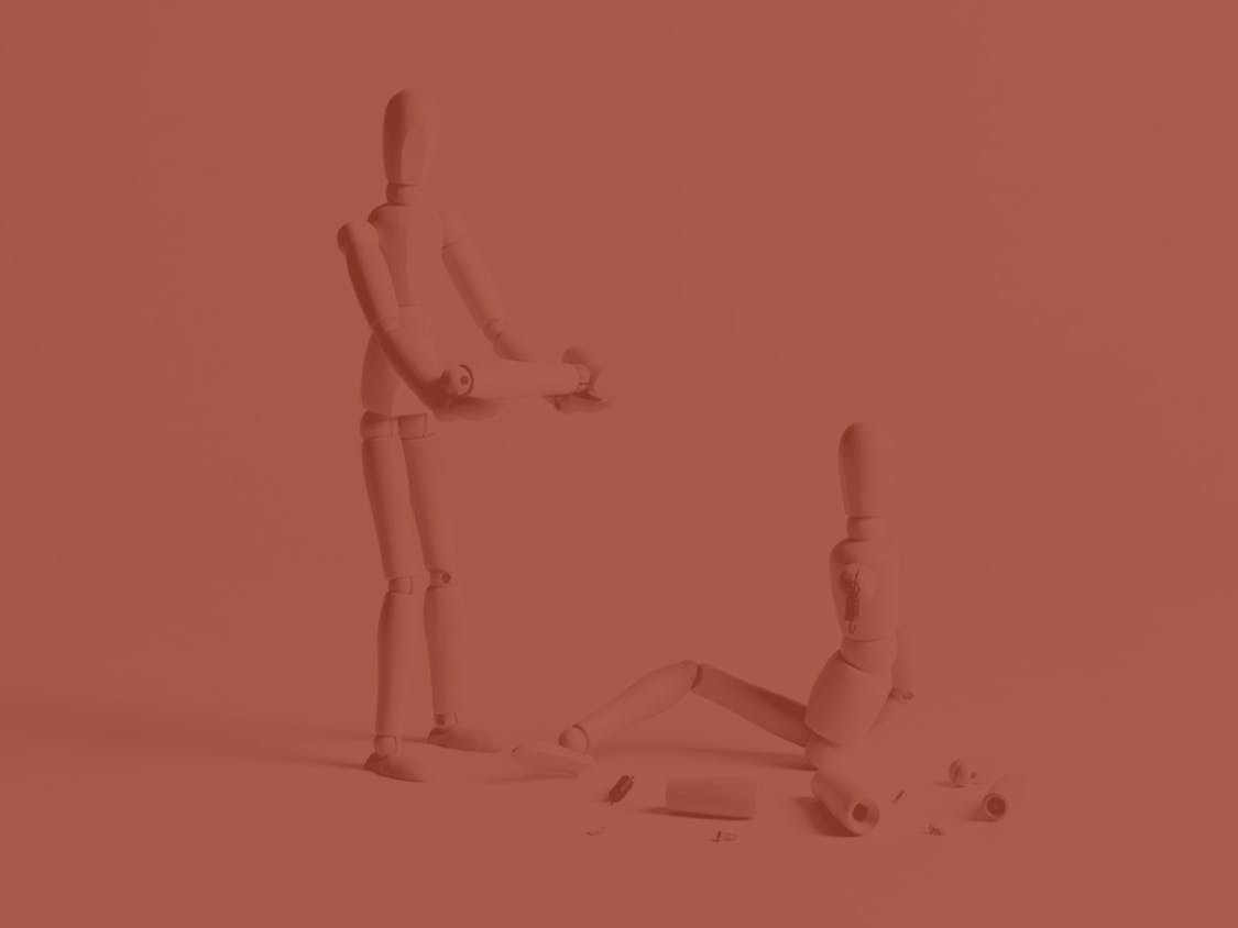 A wooden mannequin is posed to show it offering help to another wooden mannequin sitting on the ground.