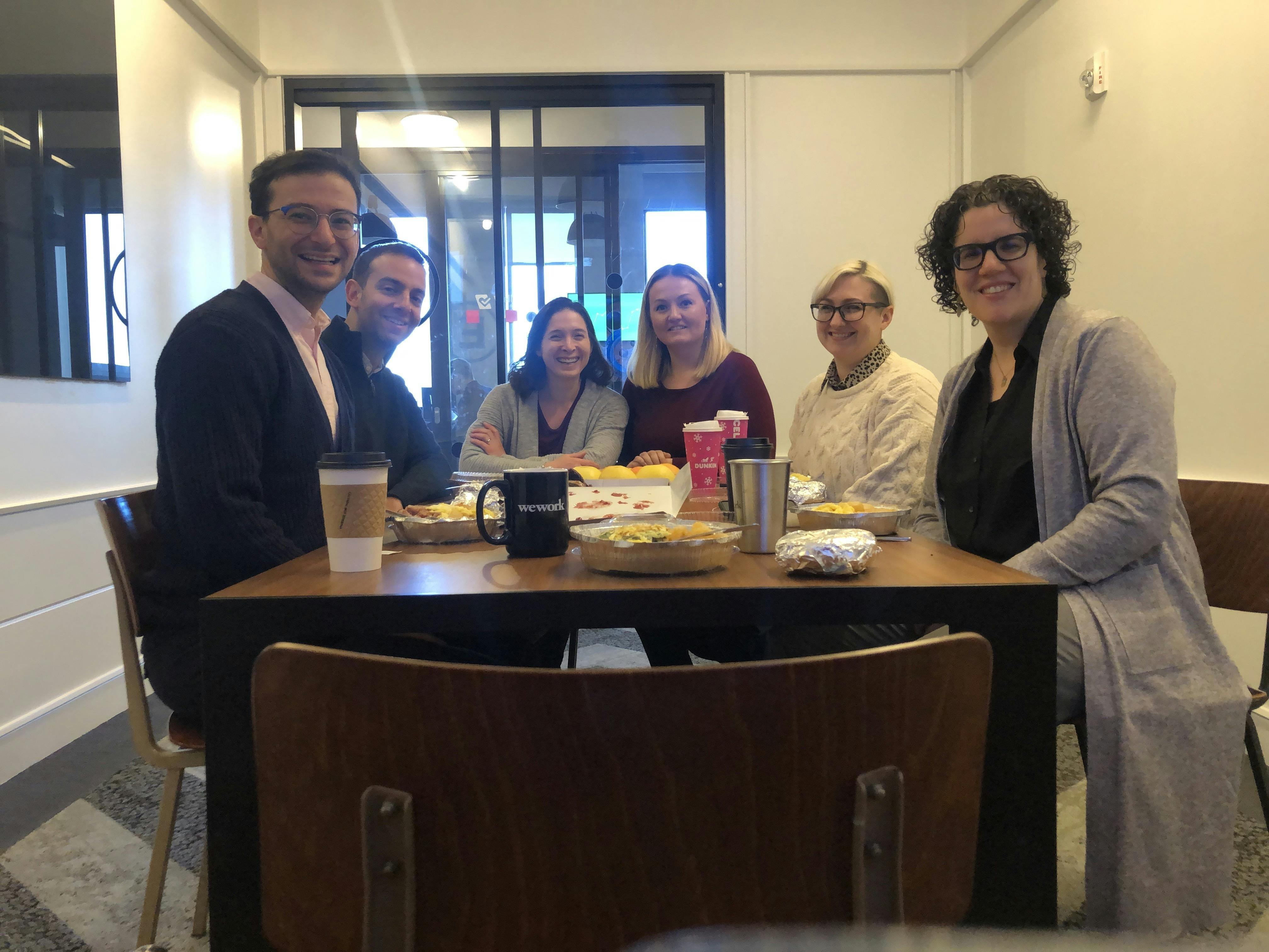 A group of six people are seated around a table, smiling, with coffee cups and food in front of them in a casual office setting